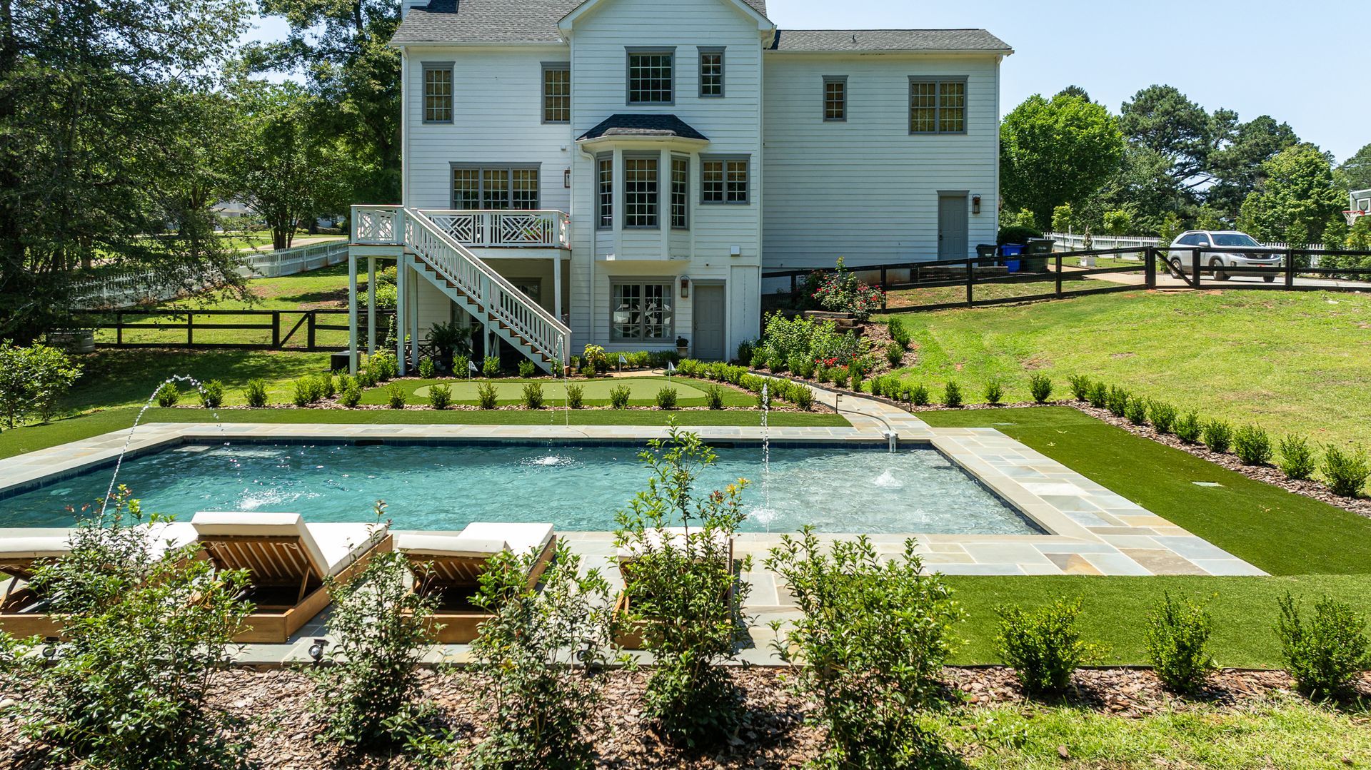 Backyard swimming pool in front of a white two-story house with lawn and trees