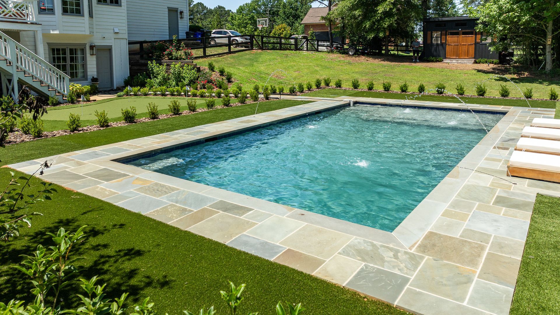 Rectangular backyard swimming pool with stone coping, surrounded by green lawn and a house in the background