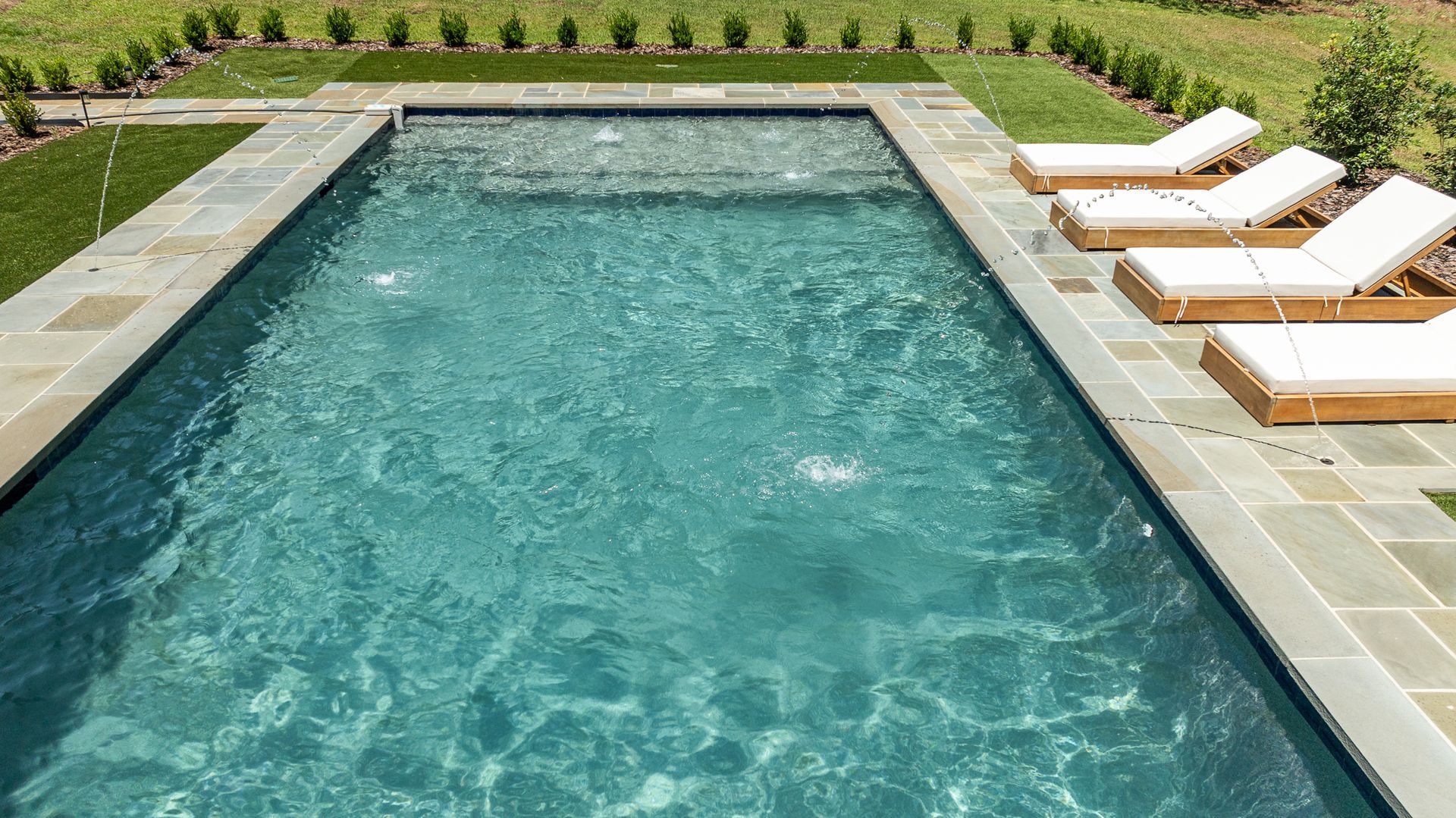 Rectangular outdoor pool with clear blue water, stone deck, and white lounge chairs on the right