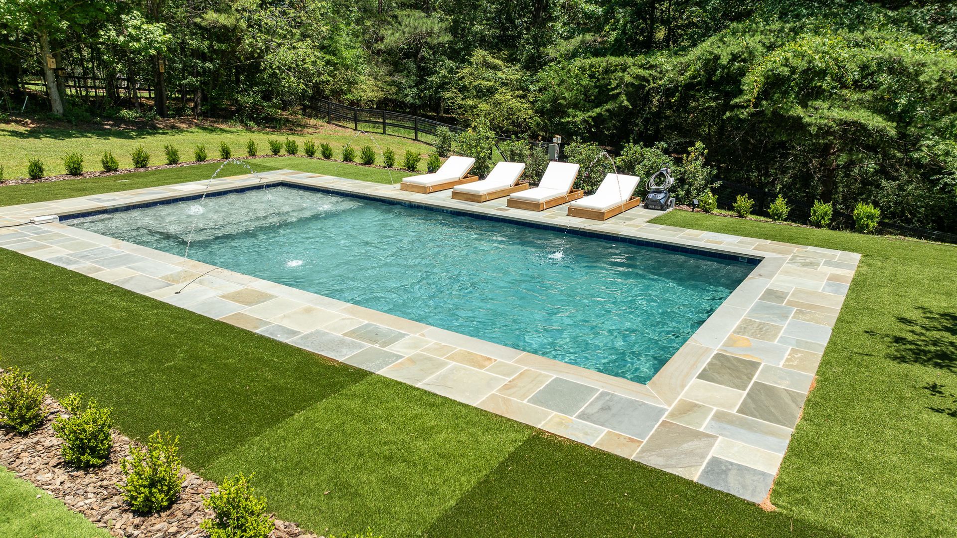 Rectangular backyard pool with stone deck and four lounge chairs, surrounded by lush green grass and trees