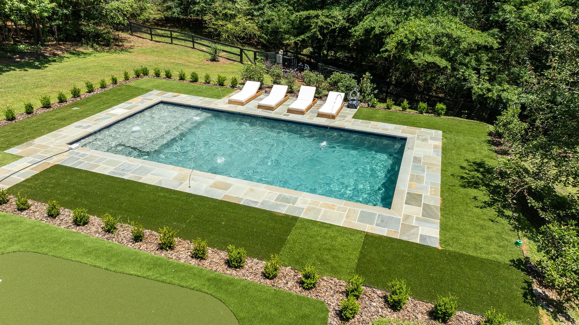 Rectangular backyard pool with stone deck, white lounge chairs, and green lawn surrounded by trees