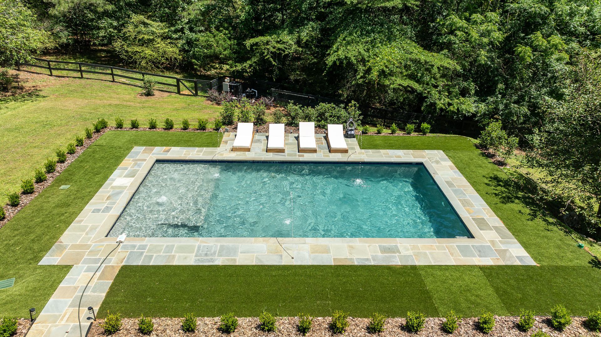 Rectangular backyard swimming pool bordered by stone, with four lounge chairs and green lawn and trees around it