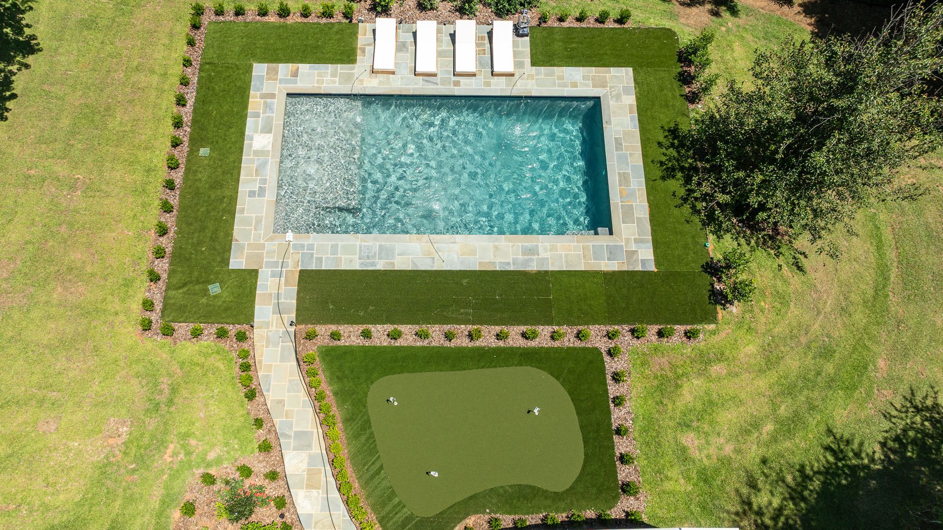 Aerial view of a backyard pool with lounge chairs, stone patio, and a small putting green.