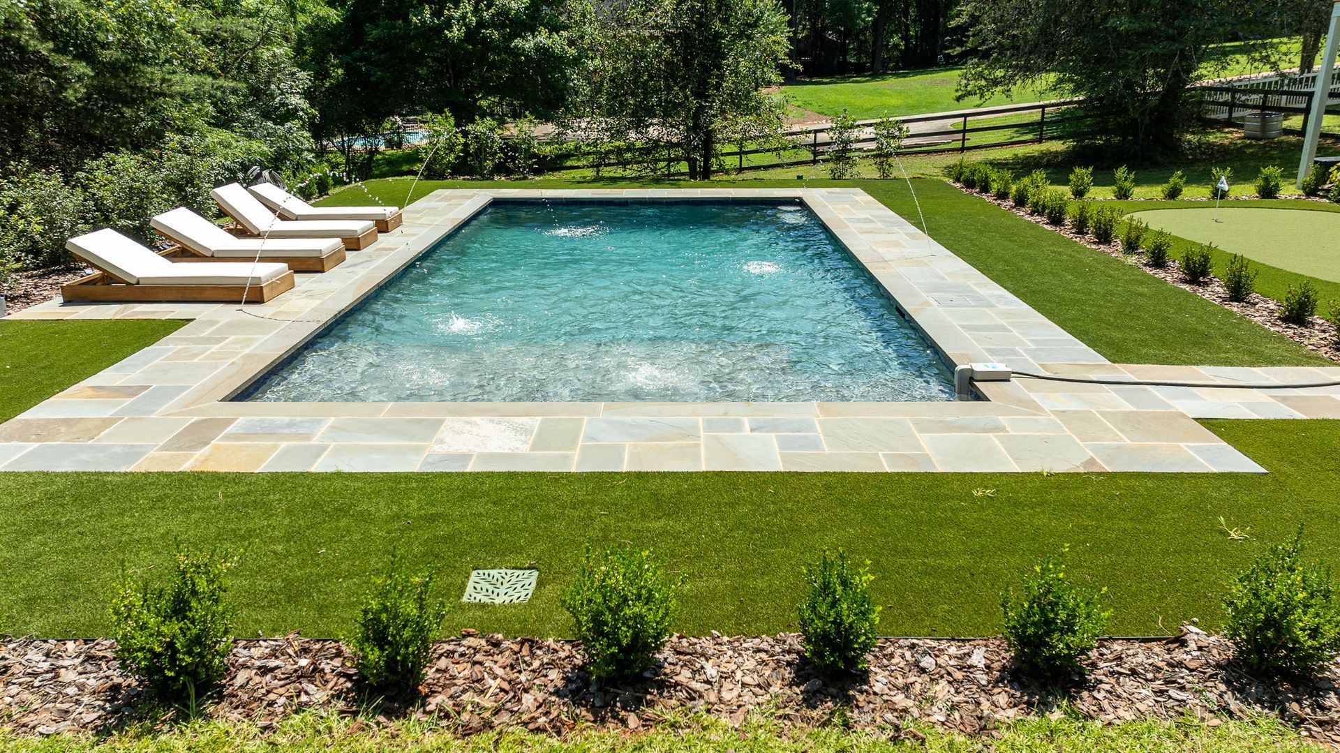 Rectangular outdoor swimming pool with stone deck, lounge chairs, and manicured green lawn