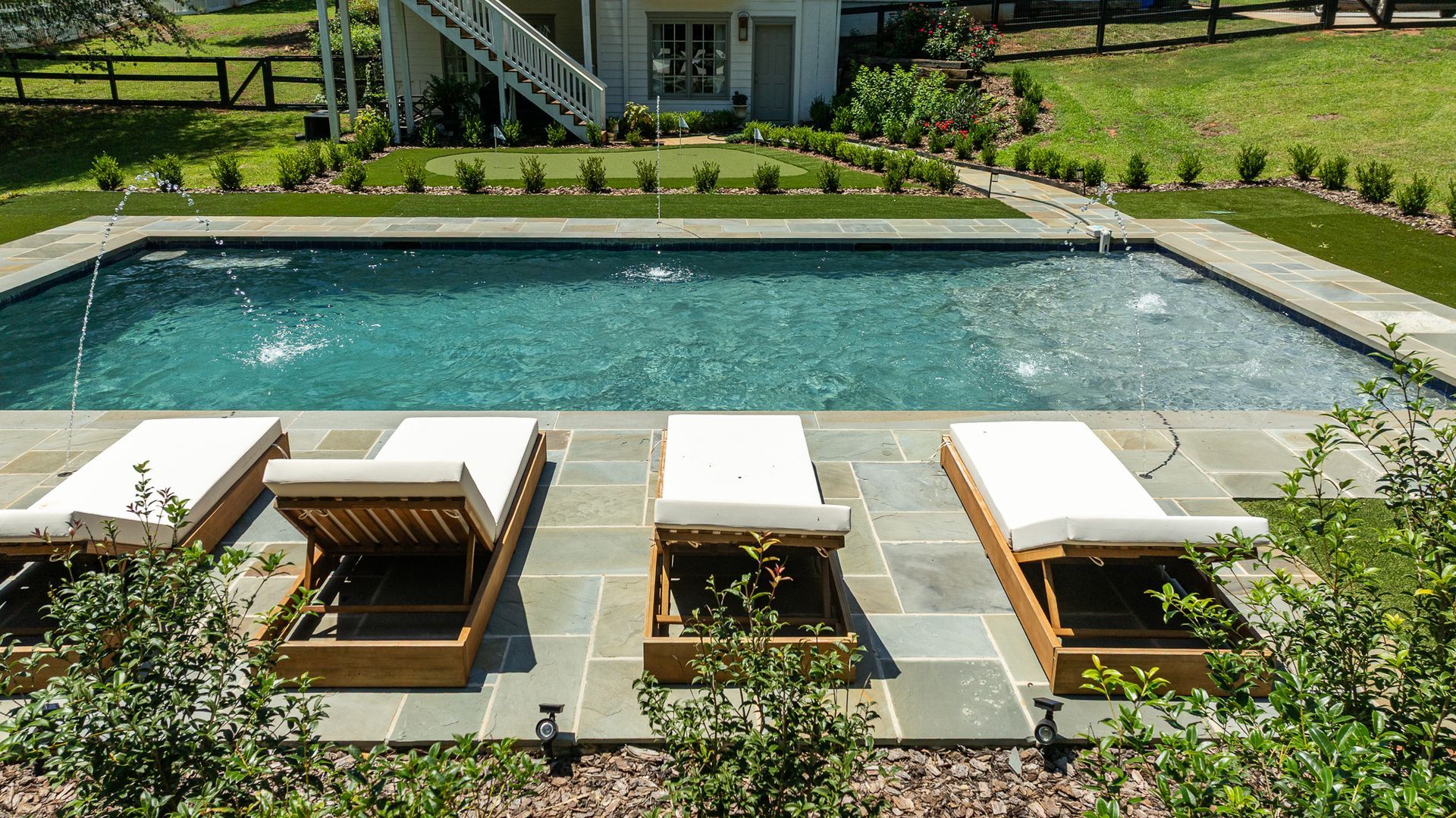 Backyard pool with white loungers and wooden daybeds on a stone patio, surrounded by greenery.