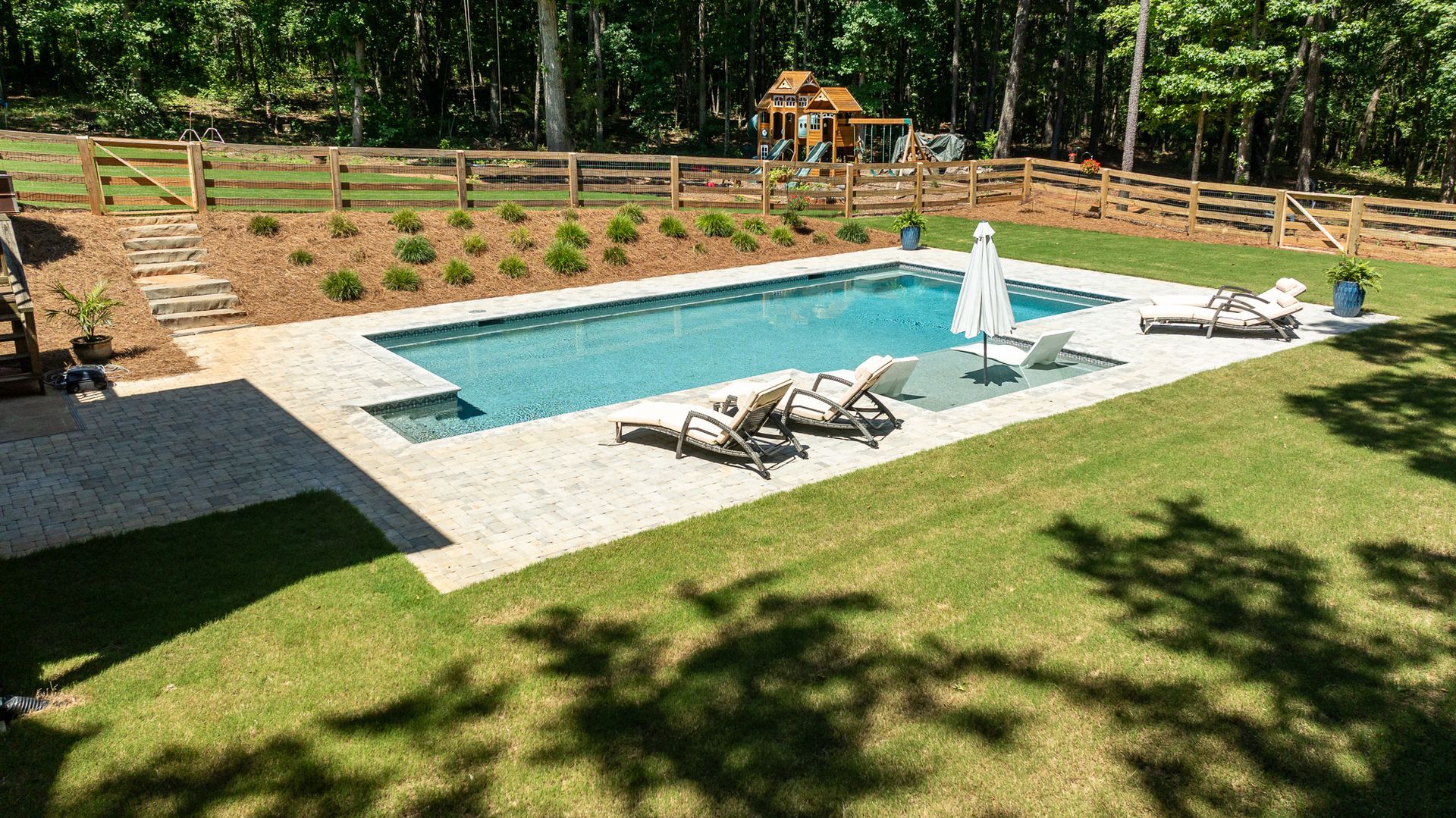 Backyard pool with lounge chairs, white umbrella, and wooden fence surrounded by trees