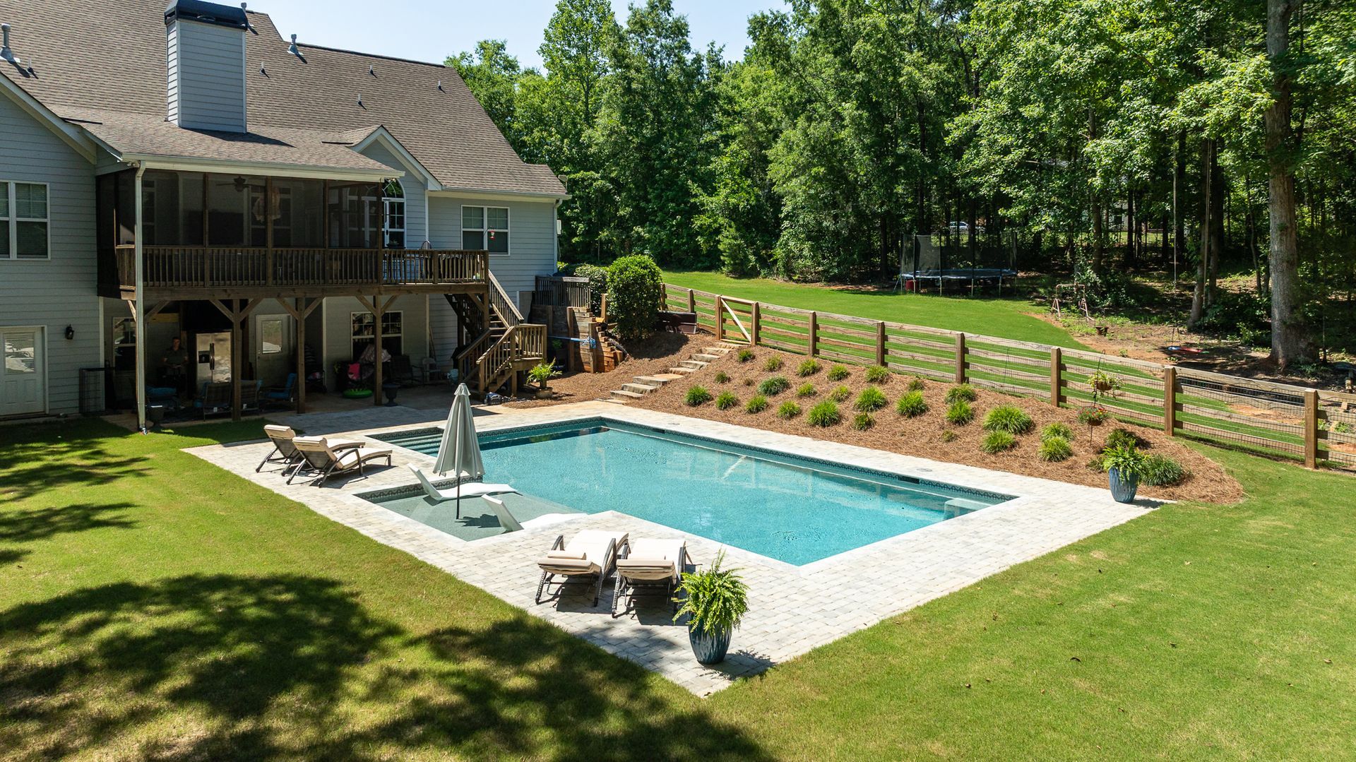 Backyard patio with a rectangular swimming pool beside a house, lounge chairs, and landscaped lawn.
