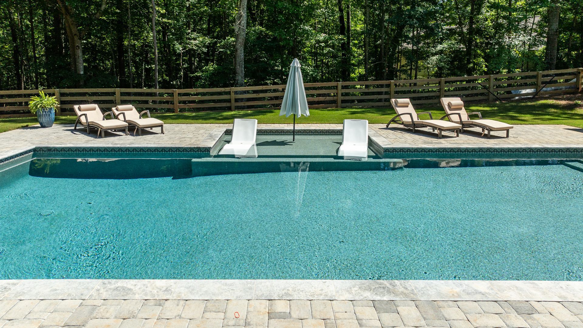 Outdoor swimming pool with lounge chairs, white umbrellas, and a wooden fence amid trees