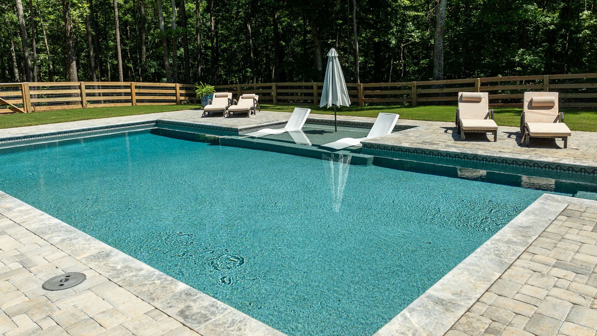 Backyard swimming pool with lounge chairs, umbrella, and a wooded fence line