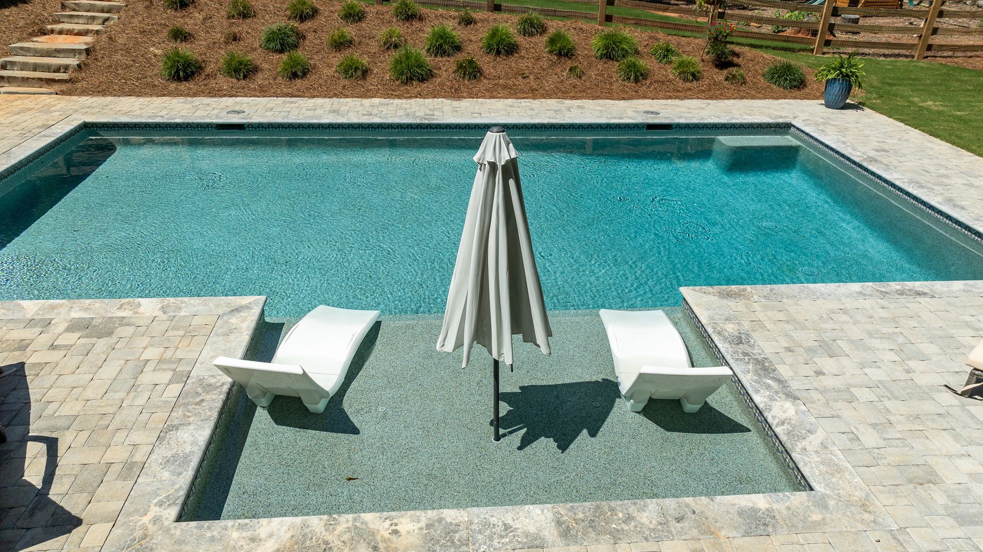 Backyard pool with two lounge chairs and a closed umbrella on a patio, landscaping in the background