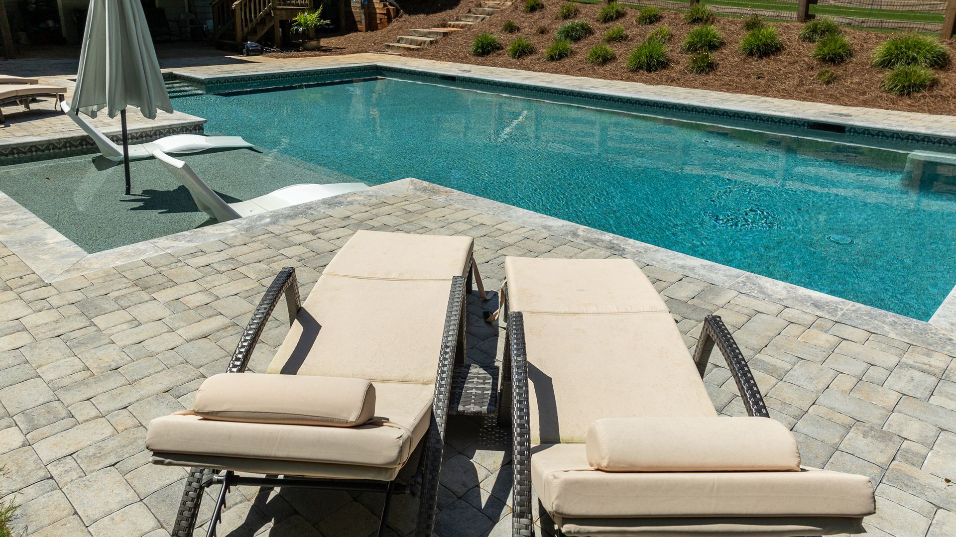 Two beige lounge chairs beside a turquoise swimming pool on a sunny patio.
