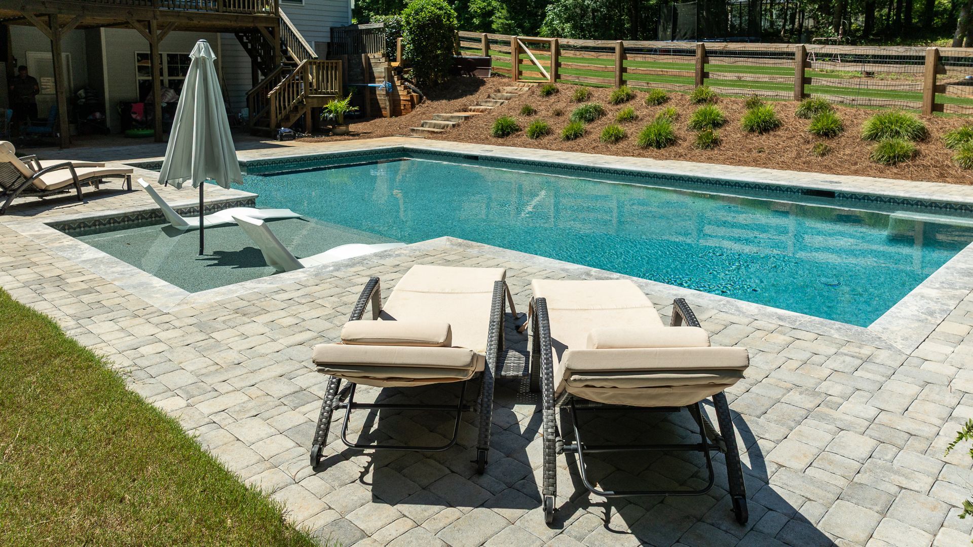 Backyard pool with two beige lounge chairs on a patio and a closed umbrella nearby