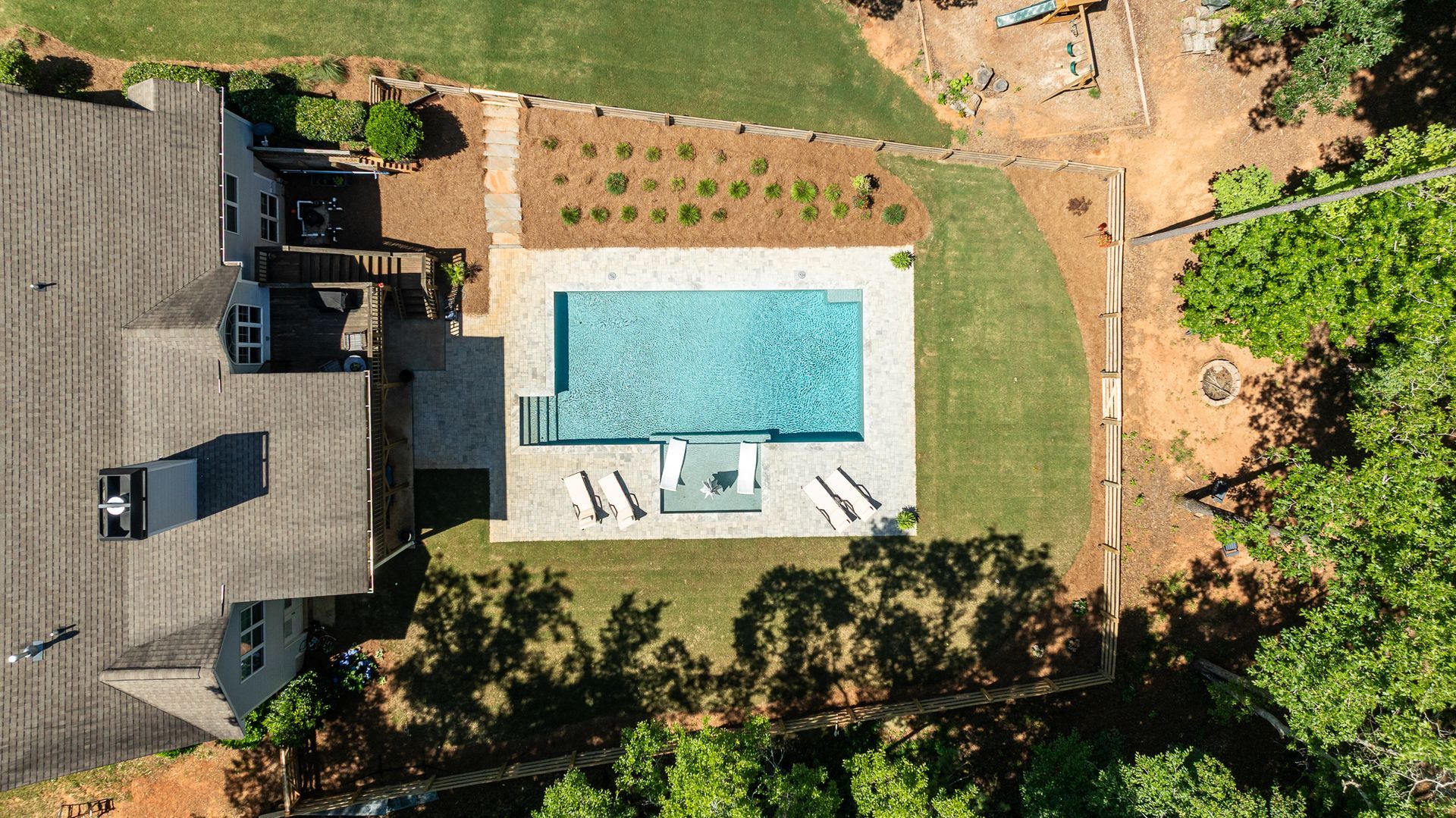 Aerial view of a backyard with a rectangular swimming pool, patio chairs, lawn, and a house roof.