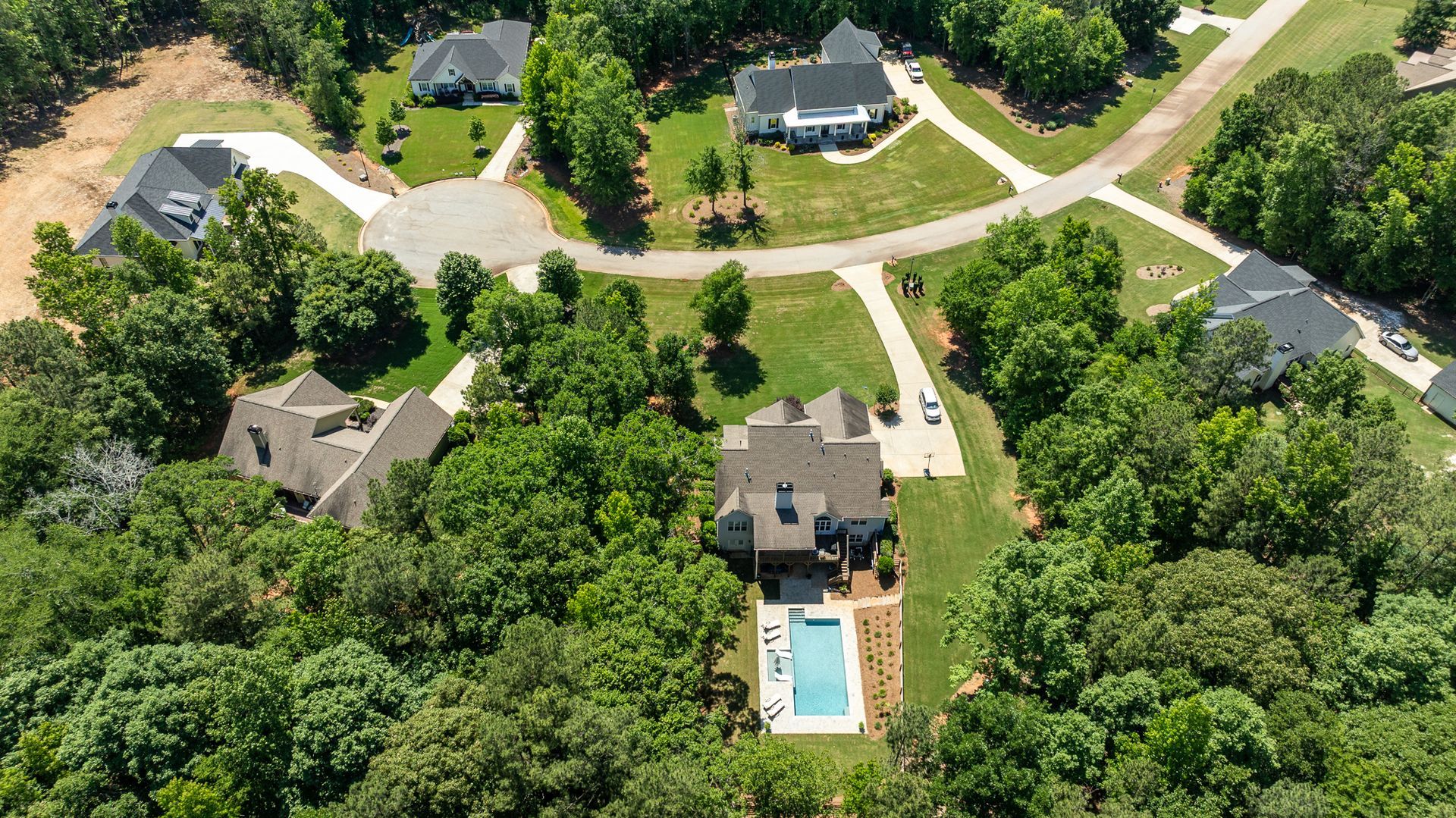 Aerial view of suburban homes along a curved road, surrounded by dense green trees, with one house and pool in the foreground