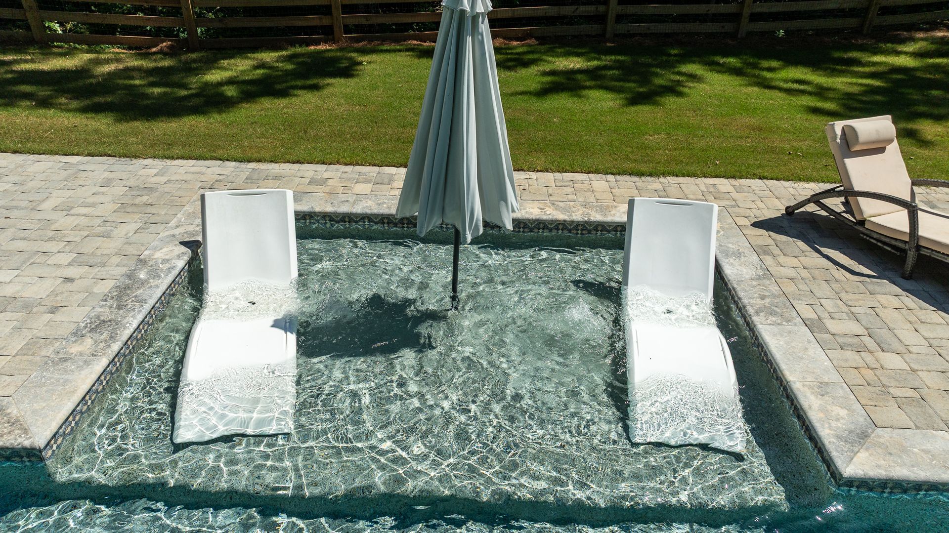 Two white lounge chairs in a small pool with a closed umbrella on a sunny patio