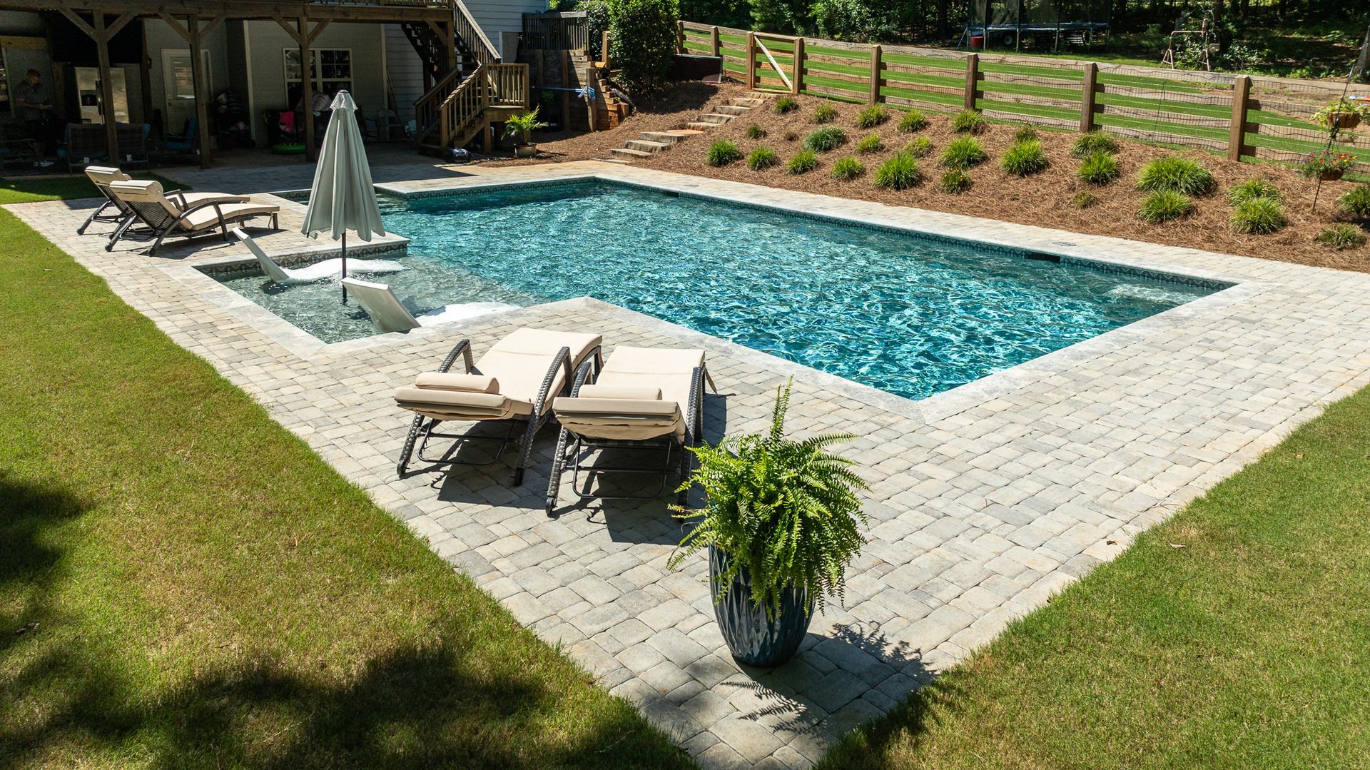 Backyard pool with lounge chairs, umbrella, and potted plant on a stone patio