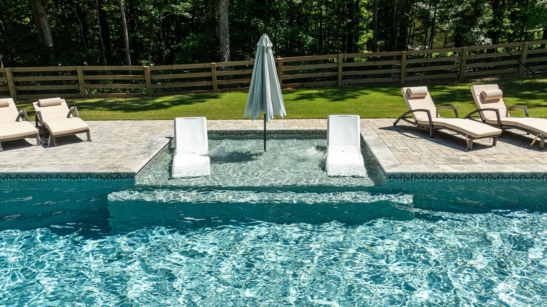 Backyard pool with two in-water loungers, umbrella, and deck chairs by a wooden fence