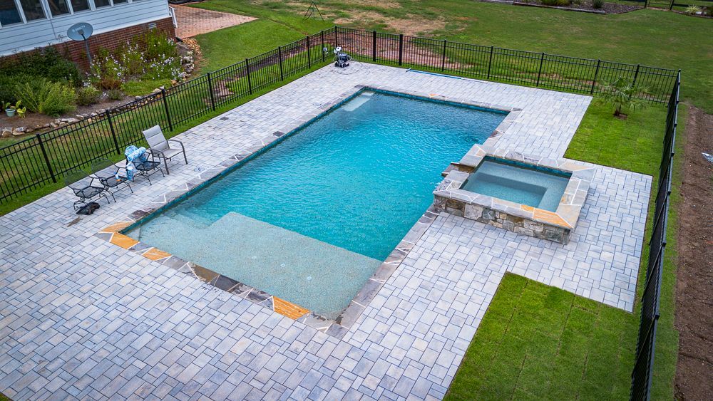 Rectangular pool with attached hot tub, surrounded by gray pavers and black fence, green grass.