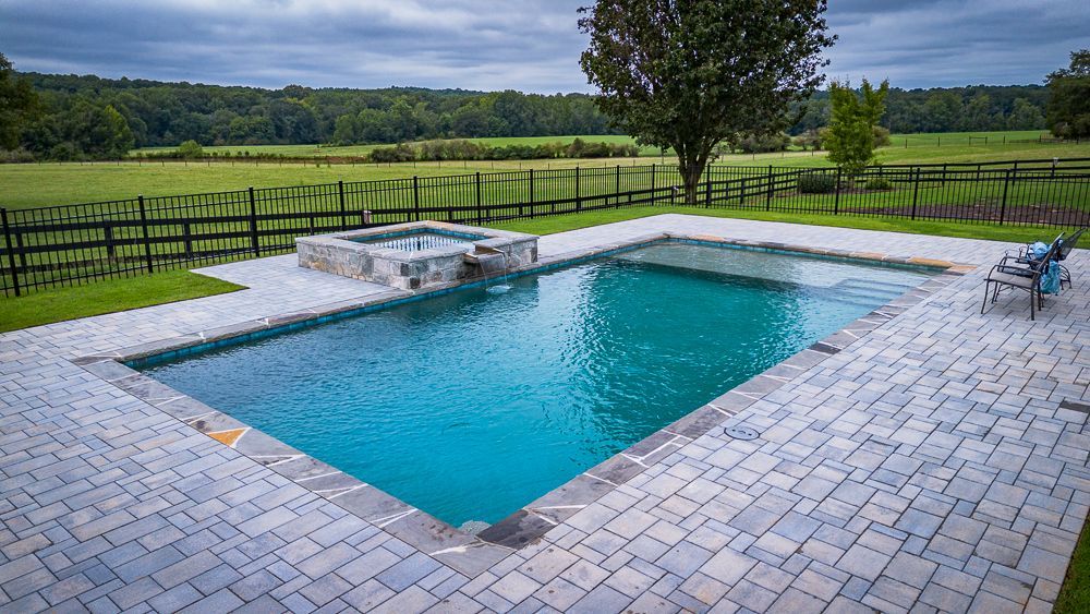 Rectangular pool with stone patio and spa overlooking a green field, black fence in the background.