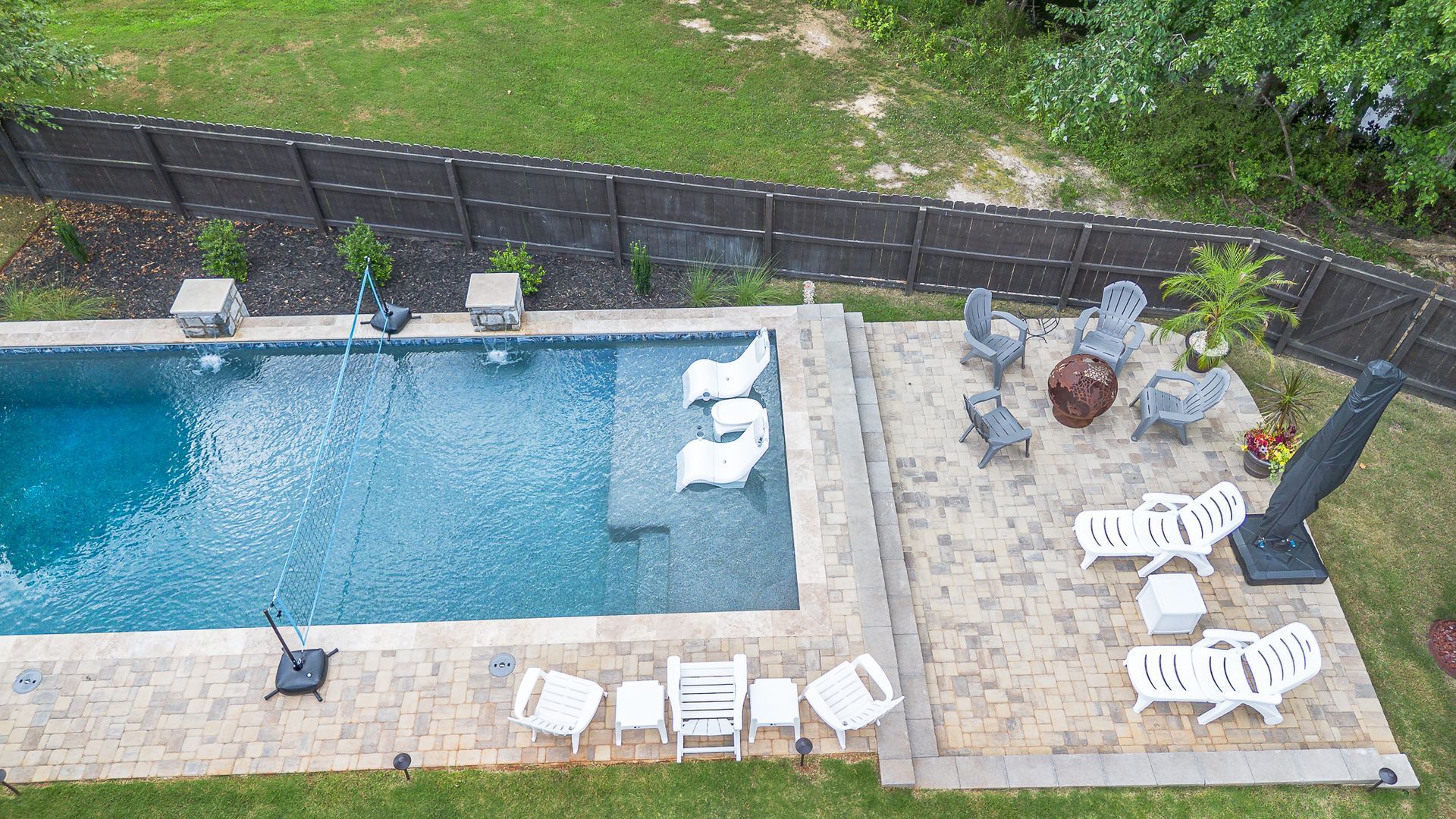 Aerial view of a backyard pool with seating areas, surrounded by a fence and grass.