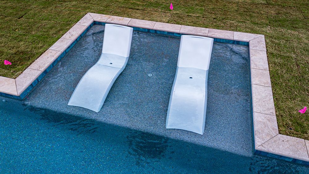 Two white chaise lounges submerged in a shallow pool section, adjacent to a larger blue pool and grassy area.