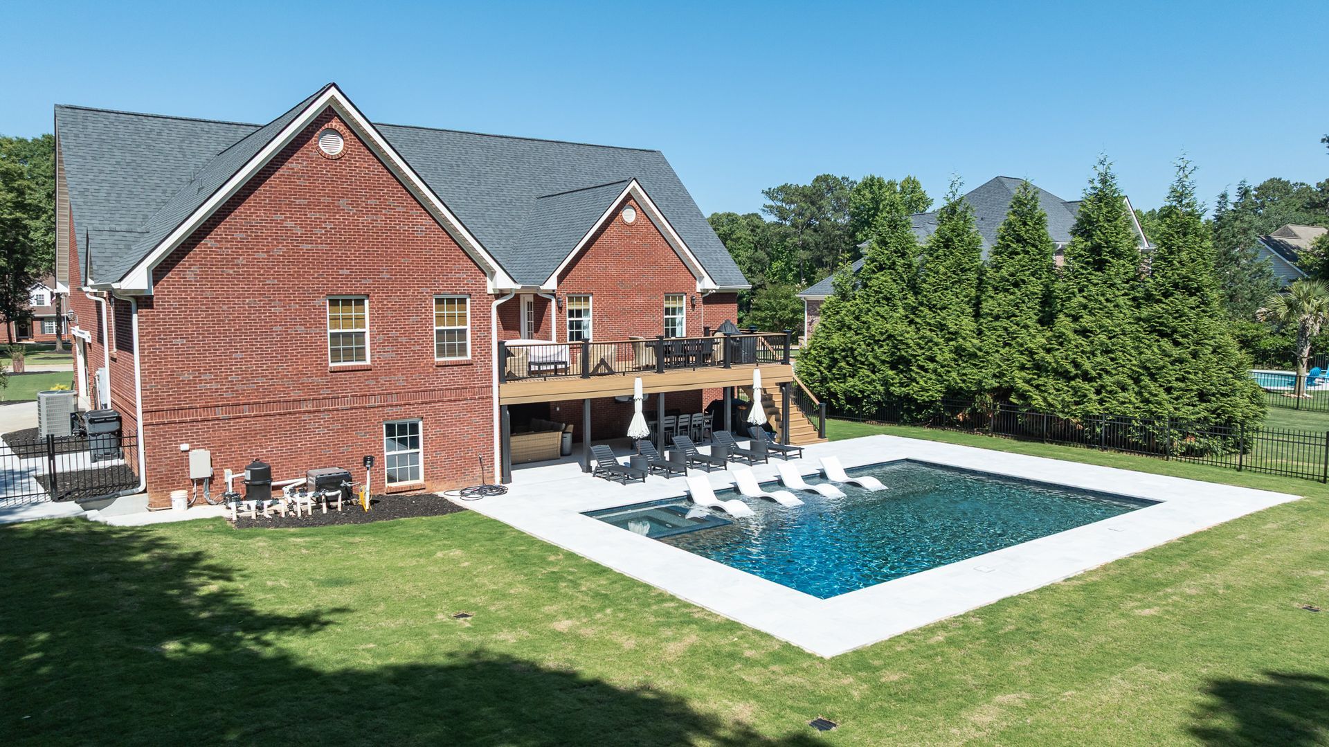 Brick house with a backyard pool, patio chairs, and green lawn under a clear blue sky