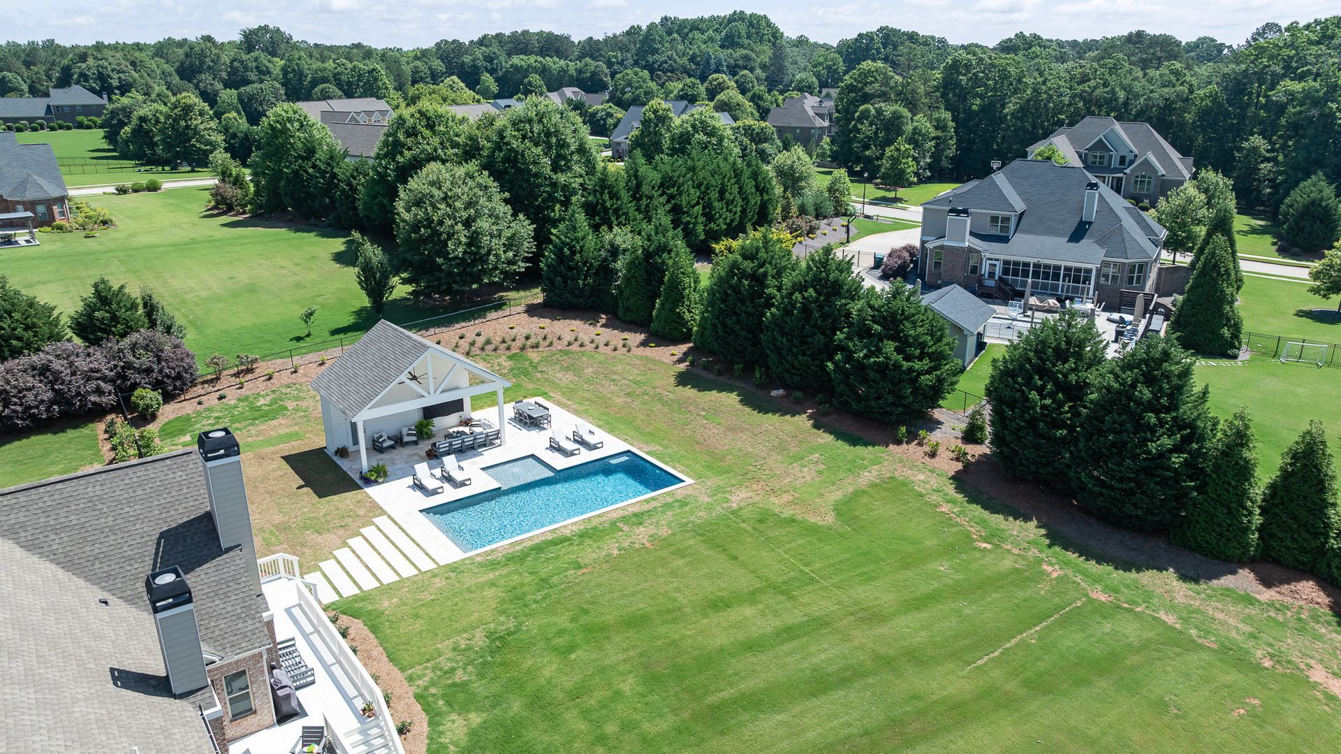 Aerial view of a suburban home with a backyard pool, patio, and large green lawn, surrounded by trees.
