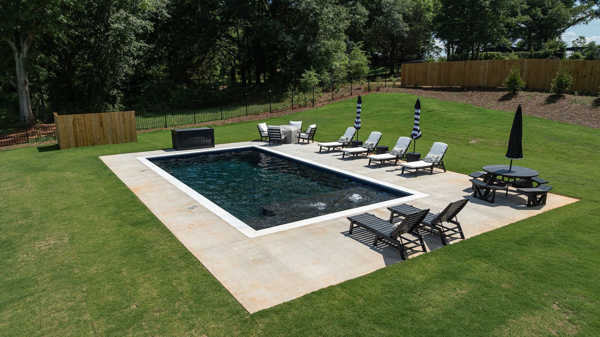 Backyard pool with white deck chairs on a green lawn, surrounded by trees and a wooden fence