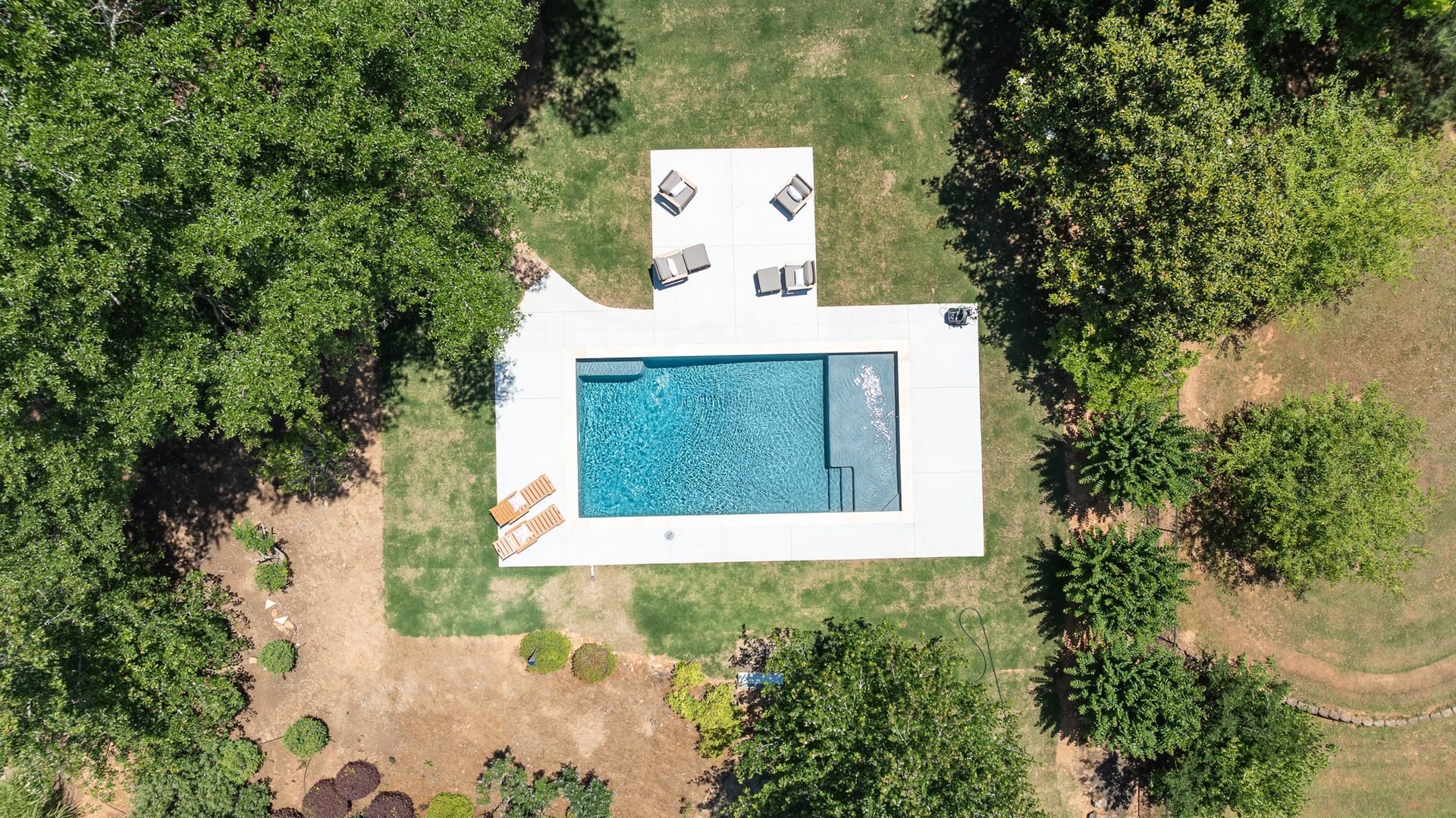 Aerial view of a backyard pool with a white deck, lounge chairs, and surrounding trees