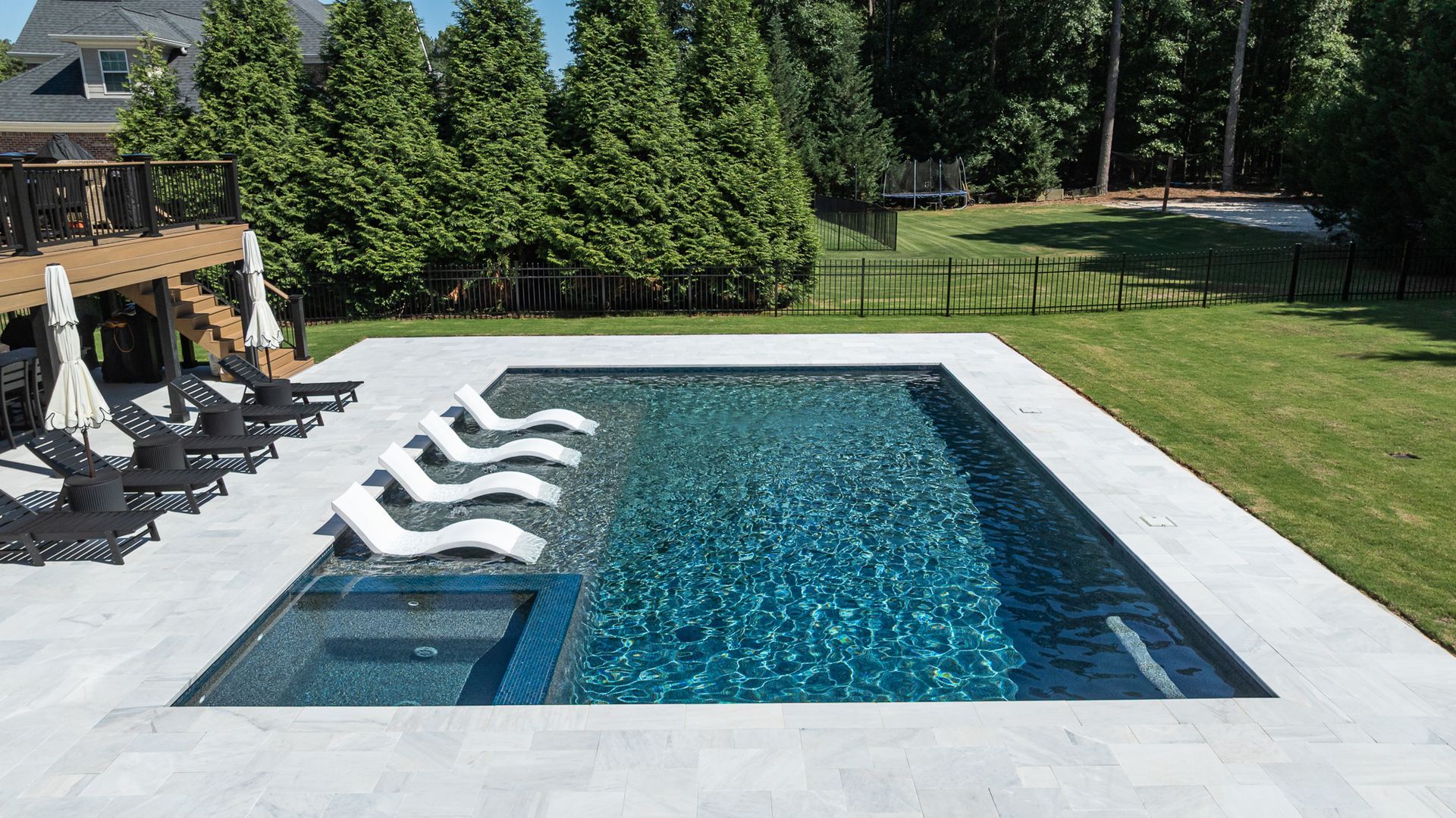 Modern backyard pool with white lounge chairs, stone deck, and green lawn trees around it