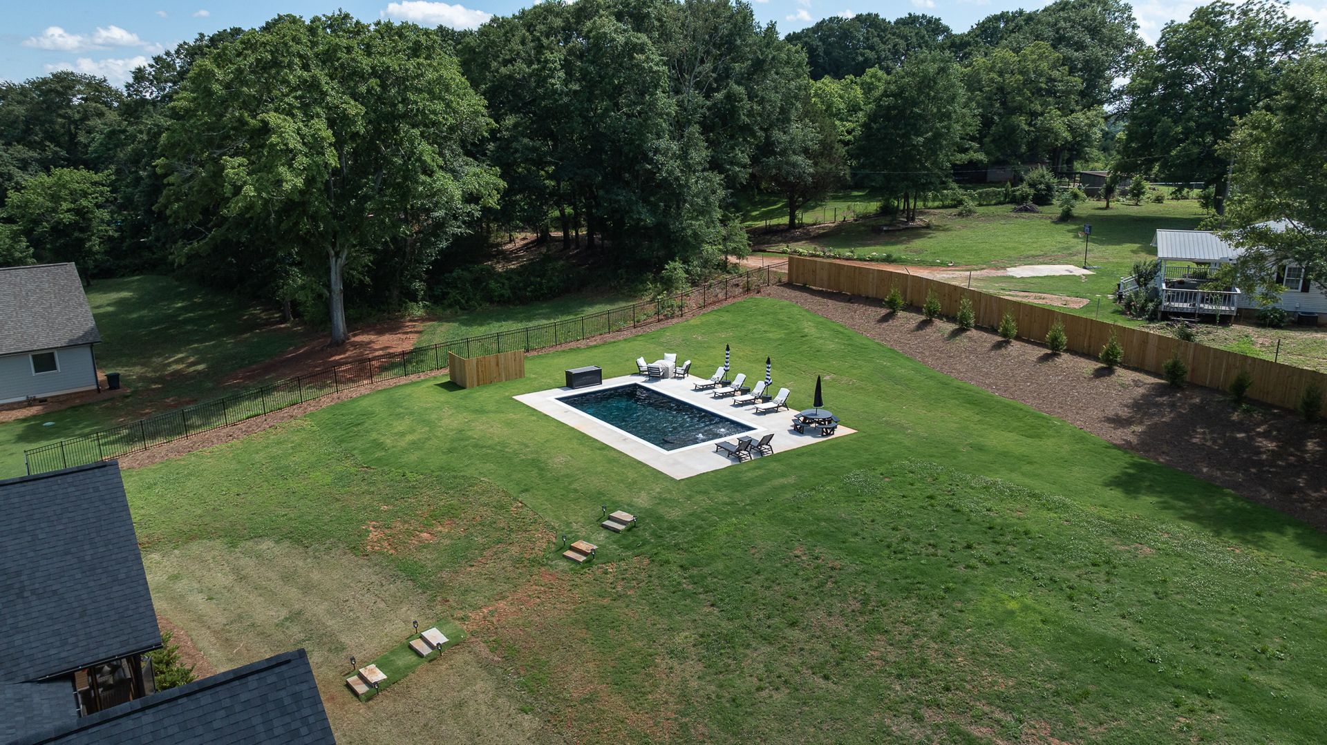 Aerial view of a backyard with a pool, lawn, and trees, bordered by a fence and houses nearby