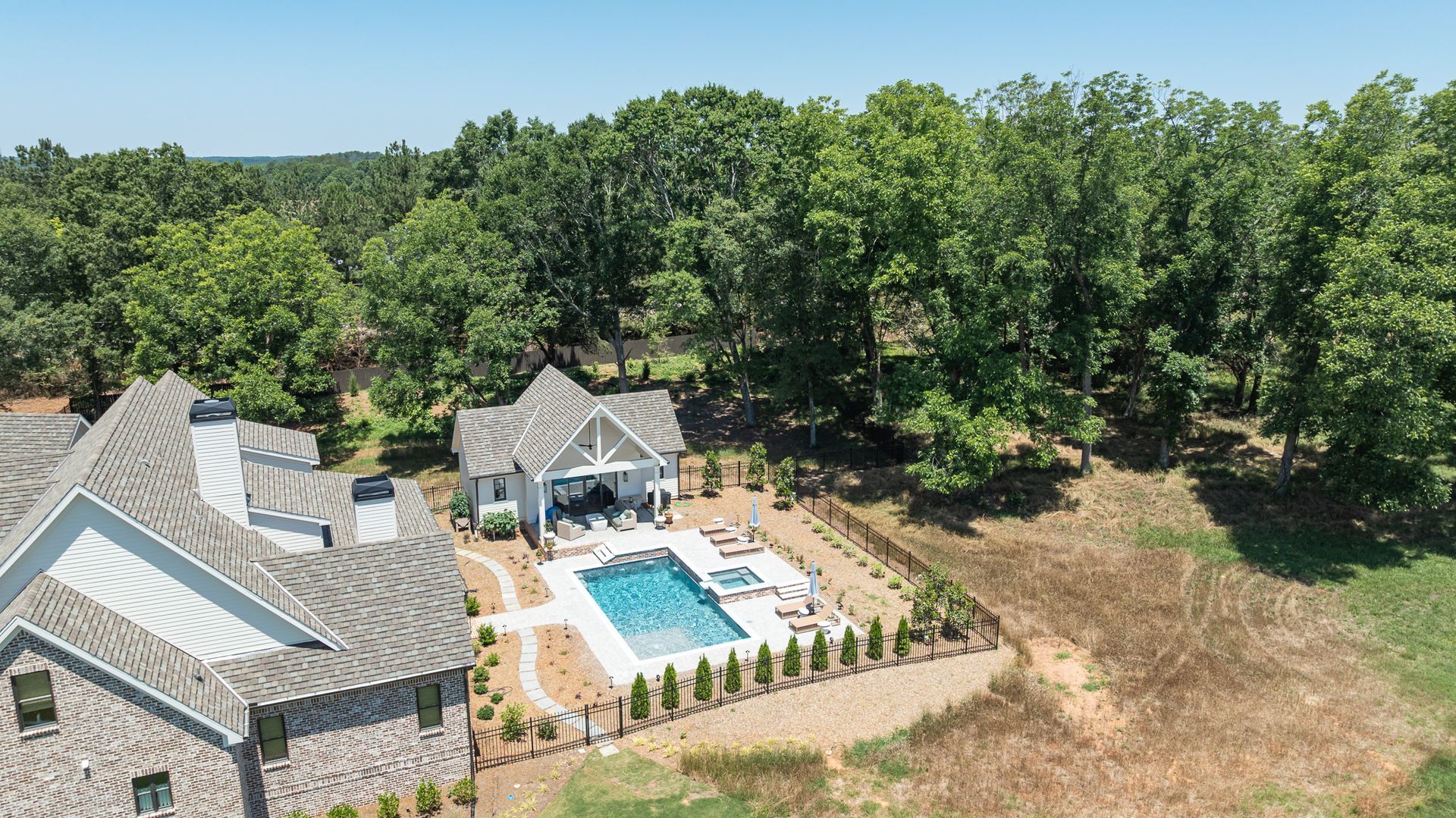Aerial view of a backyard with a swimming pool, patio, and large stone house beside a wooded area