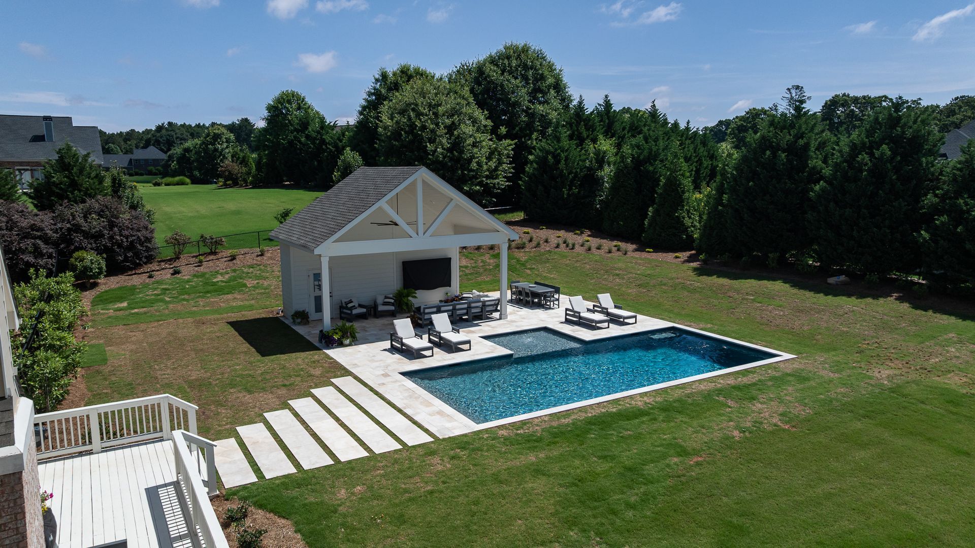 Backyard patio with blue pool, lounge chairs, and a small white pool house in a grassy yard.