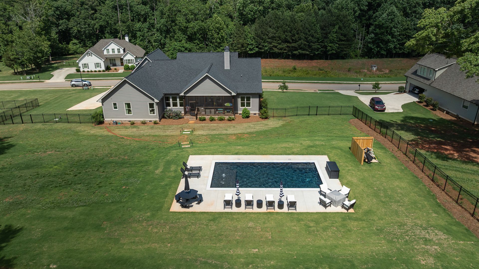 Aerial view of a suburban home with a pool, patio furniture, fenced yard, and a wooded backdrop