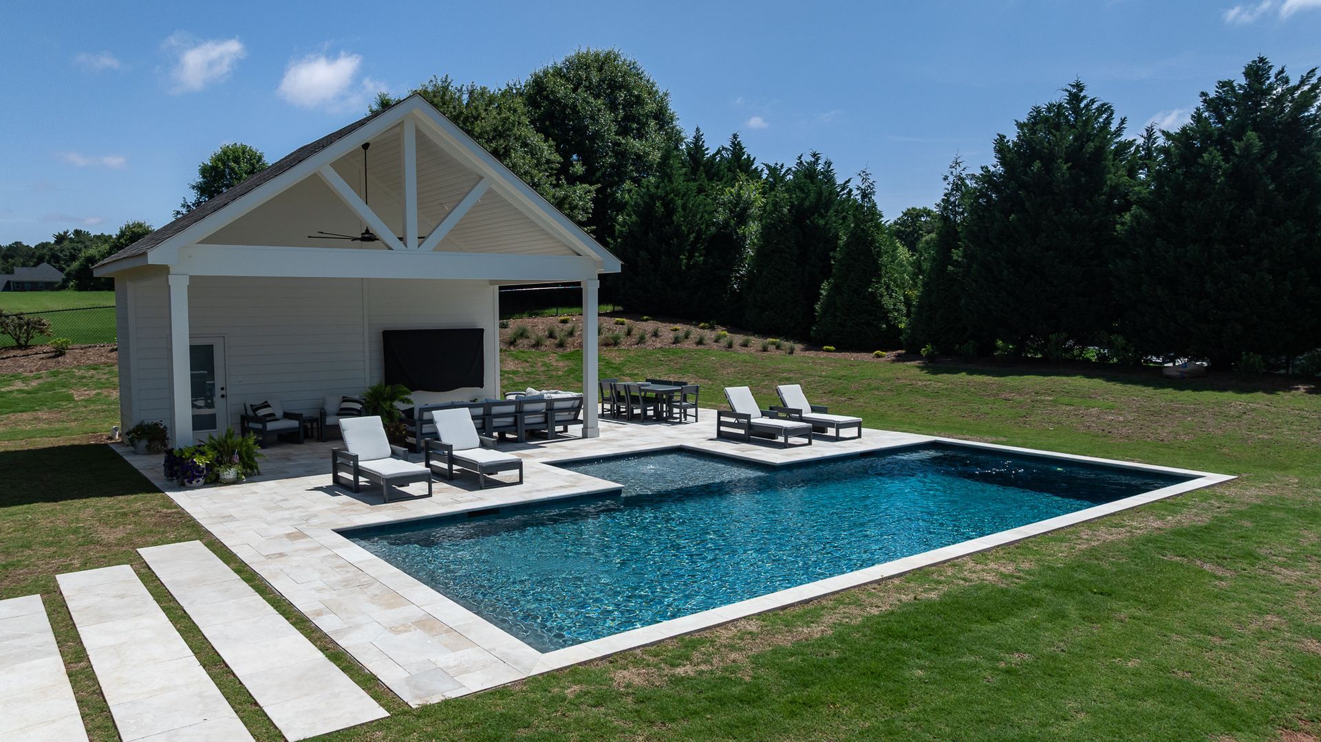 Backyard pool beside a white cabana, with lounge chairs and green lawn in bright sun