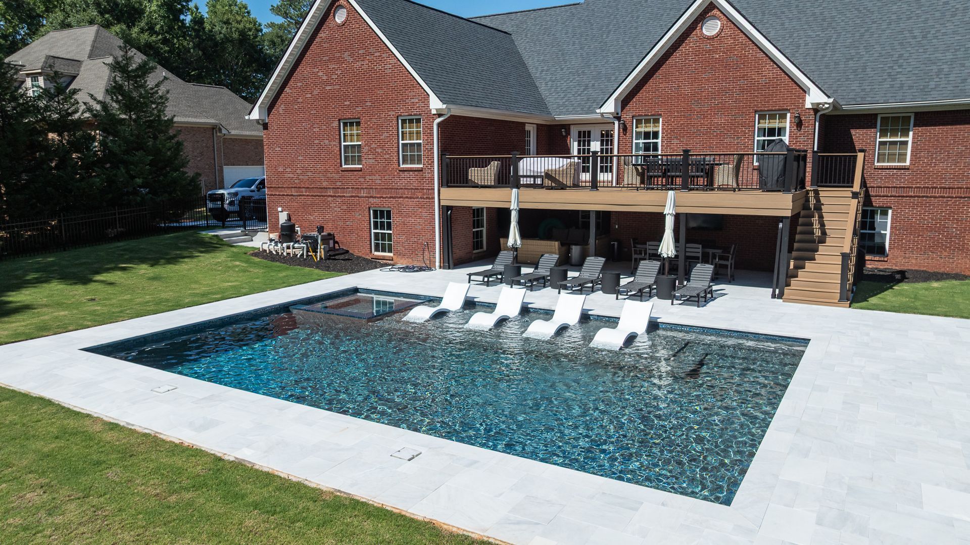 Backyard with a rectangular pool, white lounge chairs, and a brick house with a deck behind it