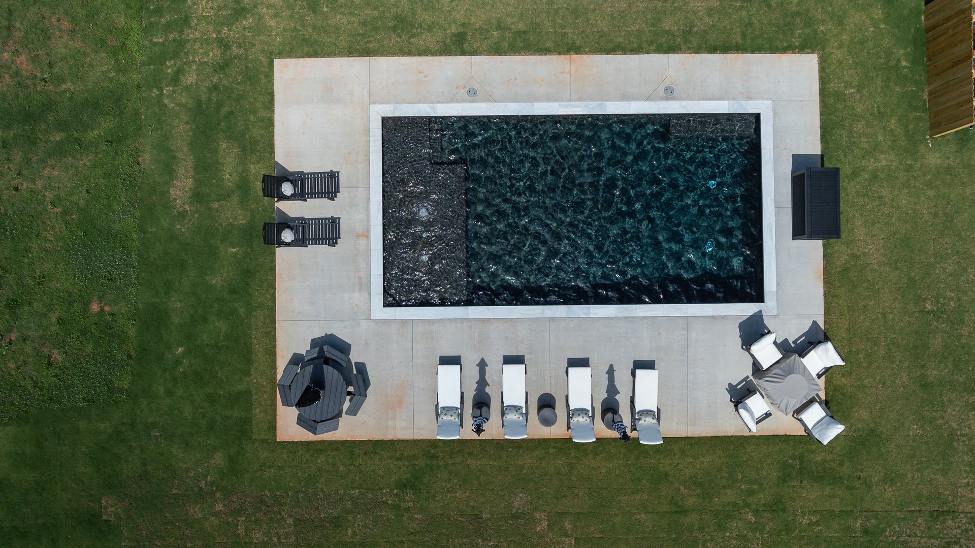 Aerial view of a rectangular swimming pool with lounge chairs and tables on a patio surrounded by grass.