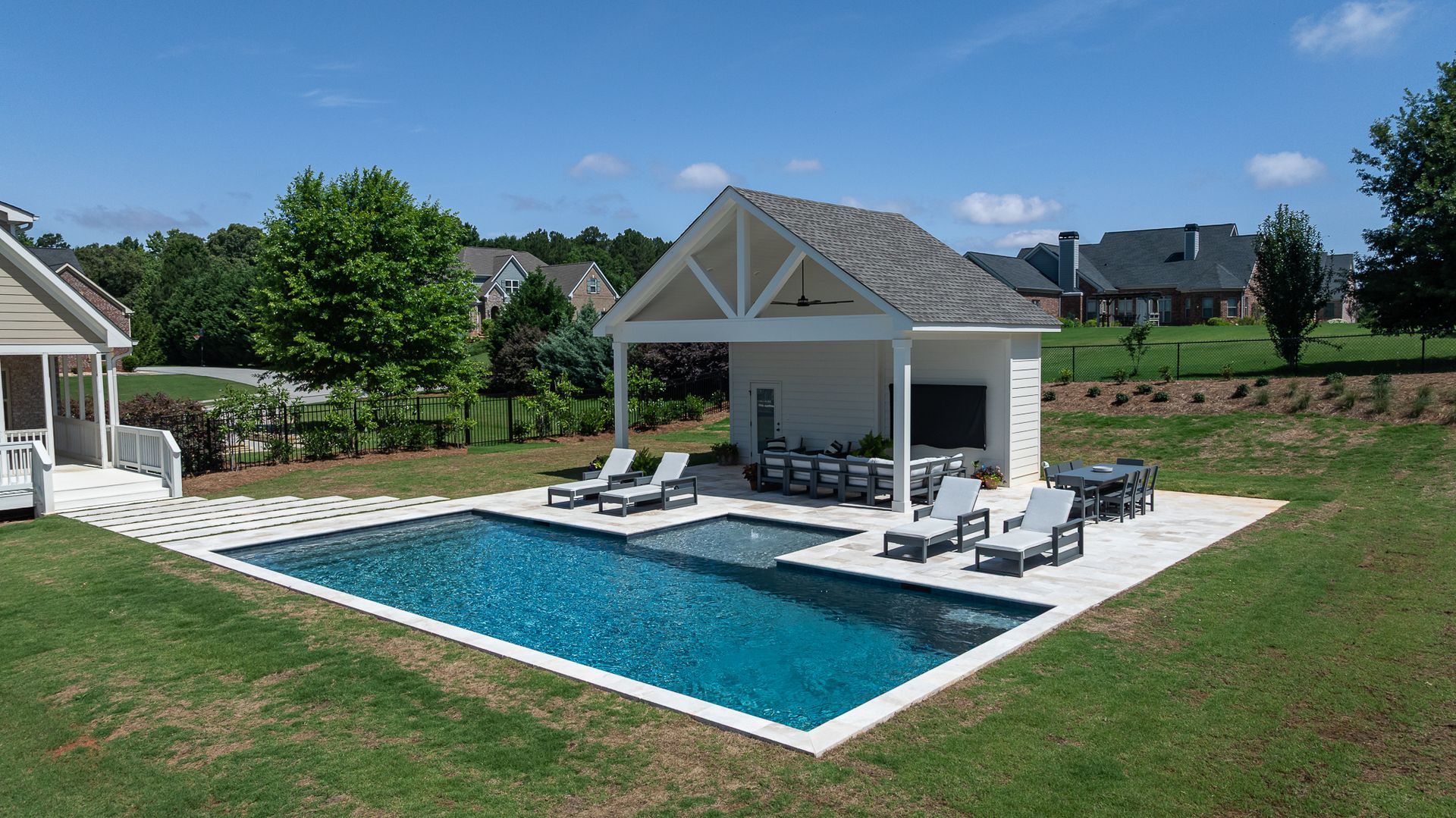 Backyard pool with a white pool house, lounge chairs, and green lawn under a sunny sky