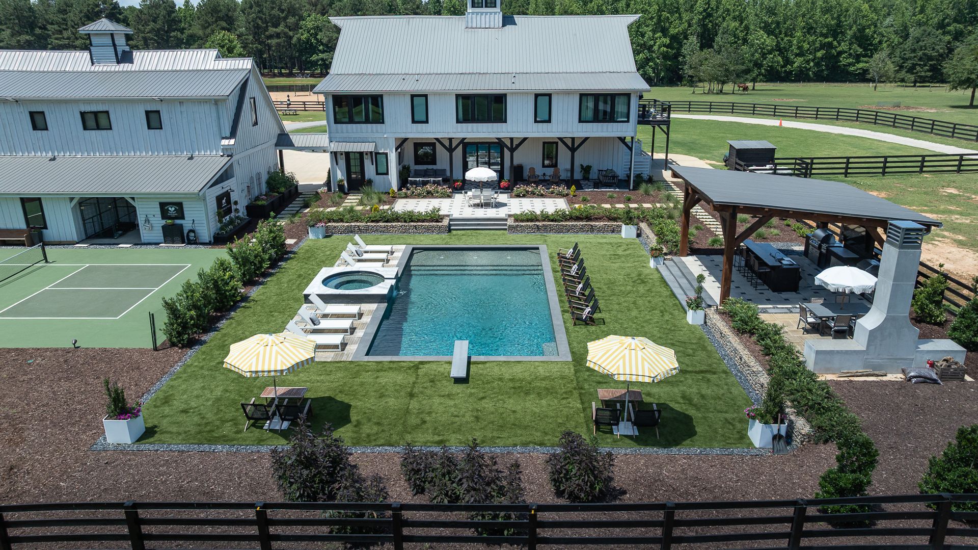 Aerial view of a backyard with a rectangular pool, loungers, umbrellas, and a covered patio beside two houses