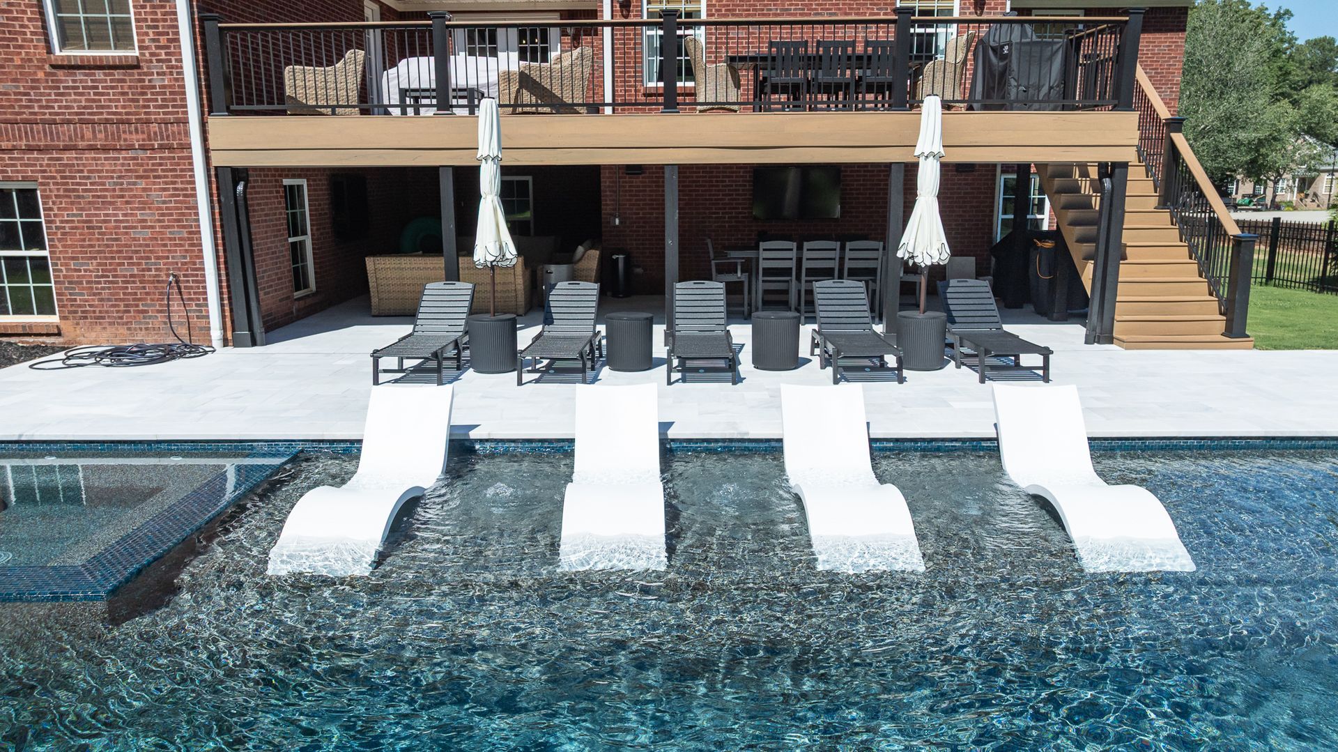 Backyard pool with four white loungers in shallow water and patio chairs under umbrellas behind them
