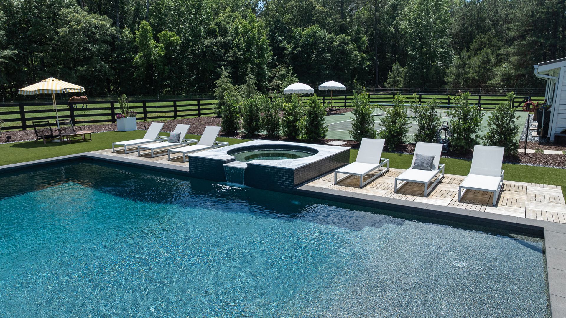 Backyard pool with lounge chairs, umbrellas, and a hot tub surrounded by trees and grass.
