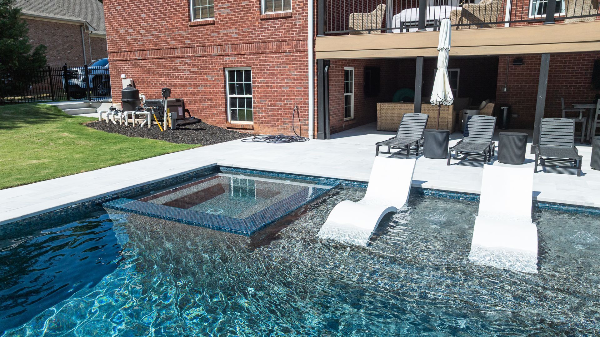Poolside patio with two white lounge chairs beside a brick building and a shallow water feature.