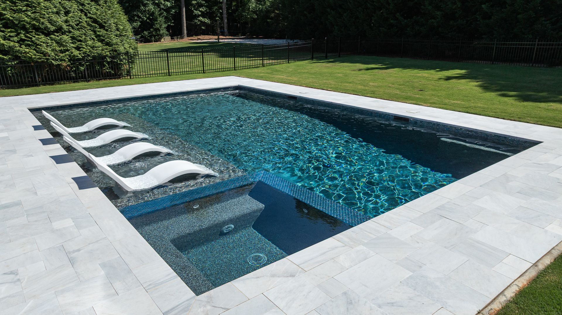 Modern backyard pool with white lounge chairs on a light stone deck and grassy yard nearby