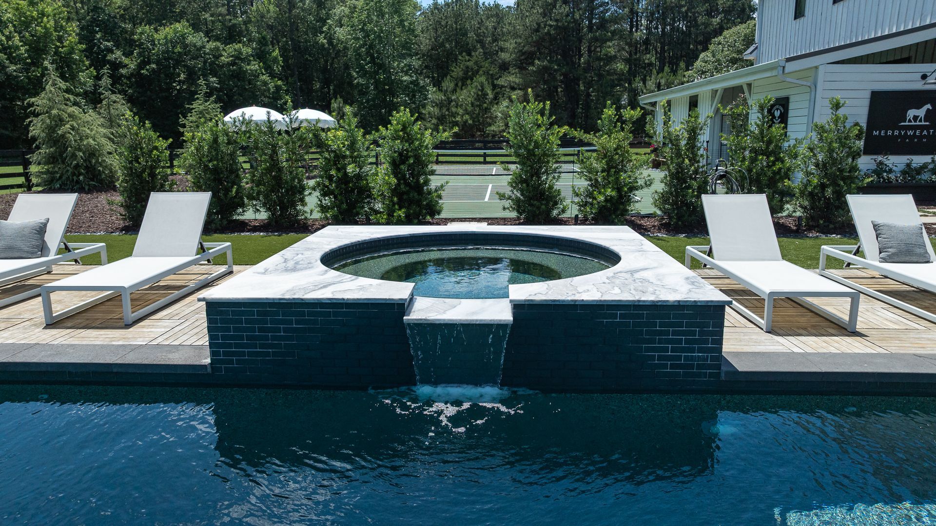 Luxurious pool deck with a round hot tub, blue water, and white lounge chairs in a sunny garden setting
