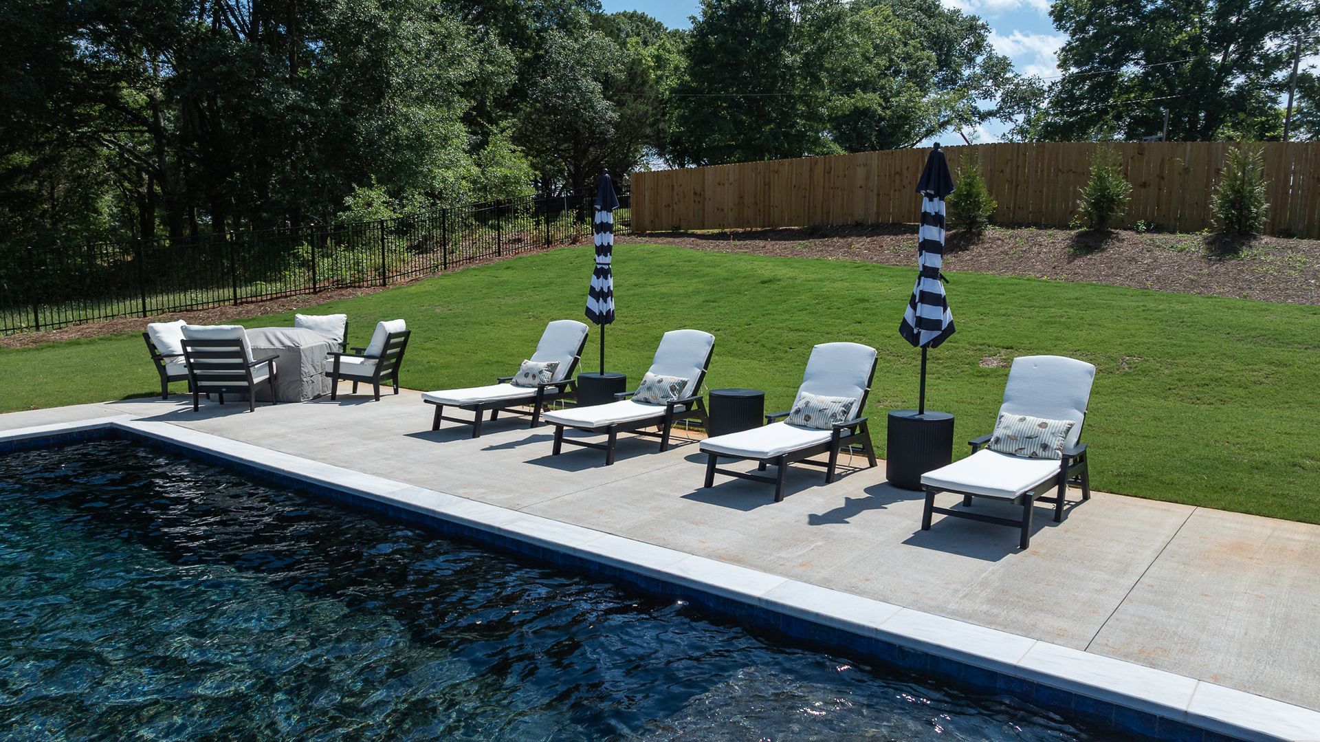 Backyard pool deck with lounge chairs, umbrellas, and a fenced lawn beyond the water