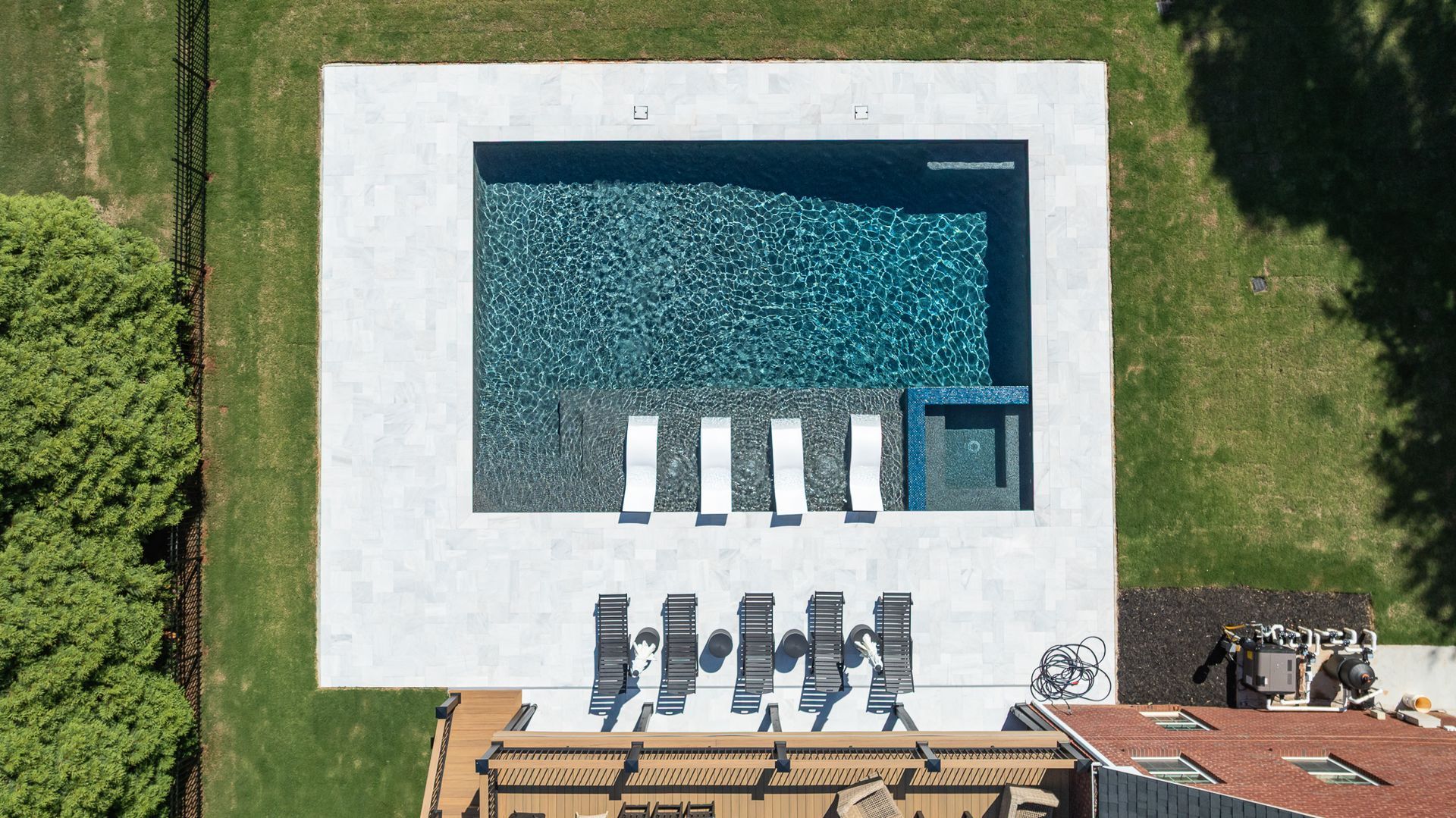 Aerial view of a rectangular swimming pool with white lounge chairs and a patio beside green grass.
