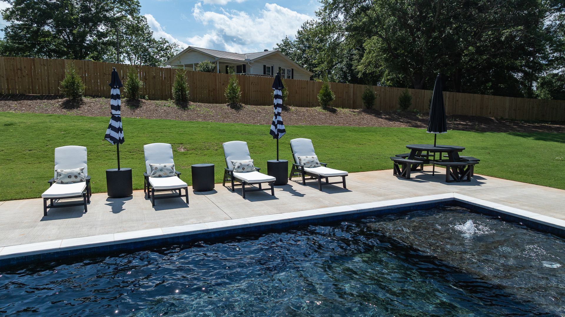 Poolside lounge chairs and umbrellas beside a blue swimming pool on a sunny lawn