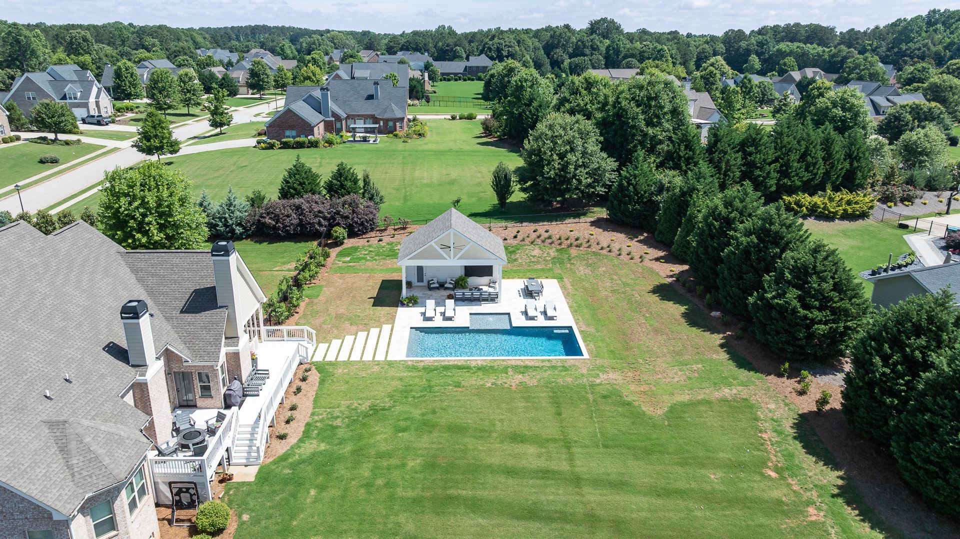 Aerial view of a suburban backyard with a house, patio, pool, and large green lawn surrounded by trees.