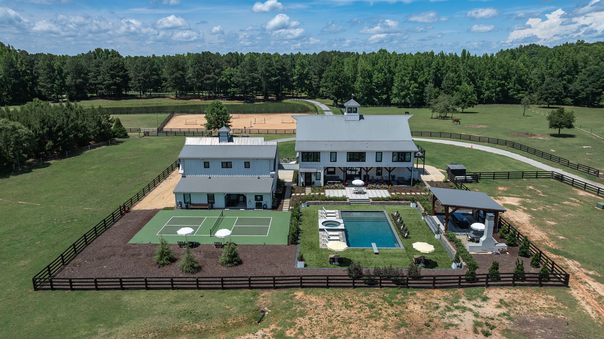 Aerial view of a fenced modern farmhouse with a pool, patio, and surrounding green fields under a blue sky