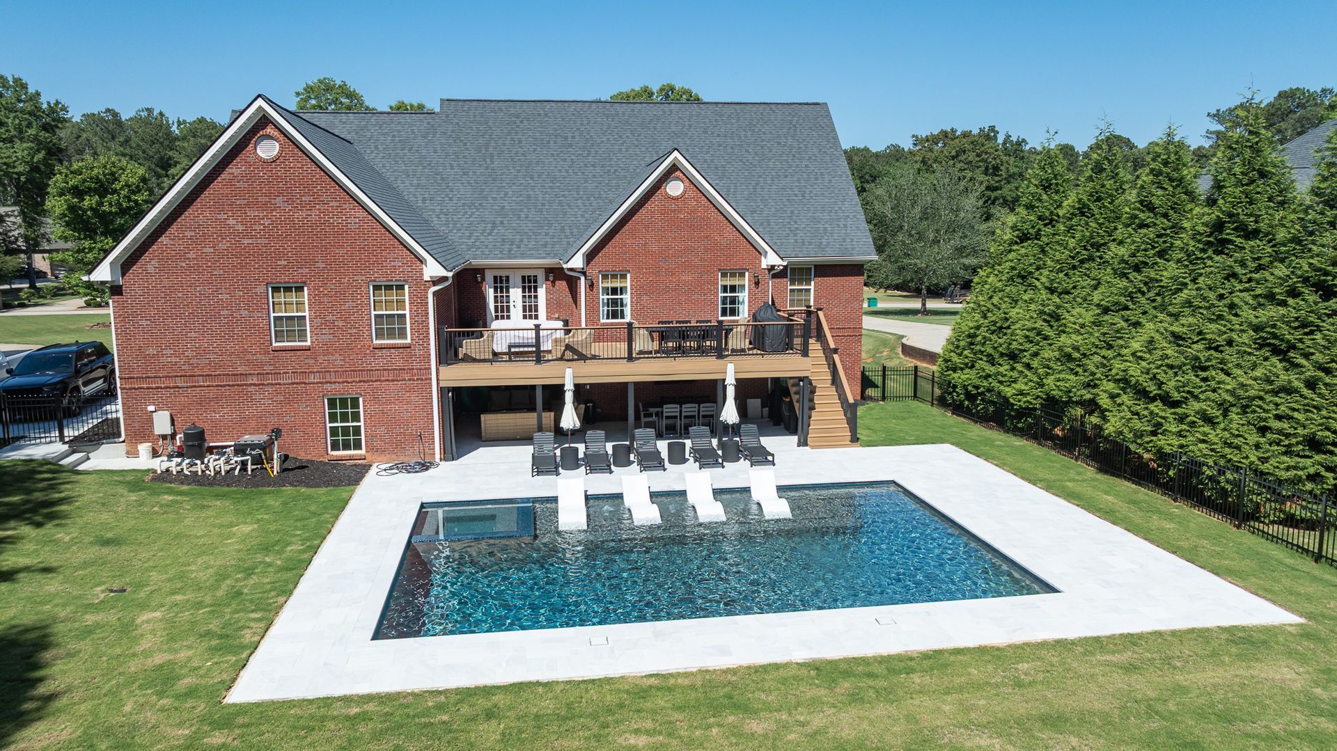 Brick house with a backyard pool and white patio, surrounded by a green lawn and trees.